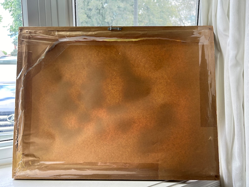 May include: The back of a framed picture with a brown paper backing and a clear plastic covering. The frame is made of wood and has a light brown finish.