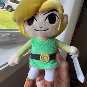 May include: A plush toy character with bright yellow hair, large black eyes, and a small stitched smile. The figure wears a green tunic with a dark brown belt featuring a yellow spiral emblem, white pants, and brown boots, holding a gray sword.