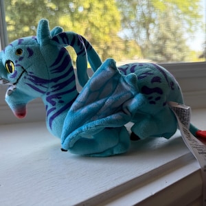 May include: A plush dragon toy featuring a bright blue body with dark purple tiger-like stripes. The creature has large yellow eyes, a small pink snout, and folded wings. A long white fabric tag with printed text is attached to the rear of the toy.