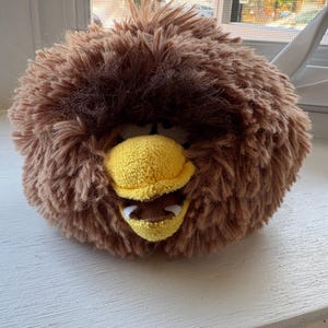 Op de afbeelding: Een pluche, ronde, bruine vogelknuffel met een gele snavel en open mond. Het speelgoed heeft een harige, getextureerde uitstraling, met donkerbruine accenten rond de ogen. De vogel zit op een wit oppervlak.