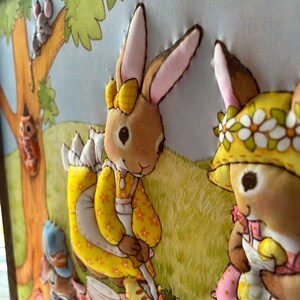 May include: A framed fabric wall art piece depicting two bunnies having a picnic in a forest setting. The bunnies are wearing dresses and have floral headbands. The scene includes a picnic basket, a pie, and a tree with a squirrel.
