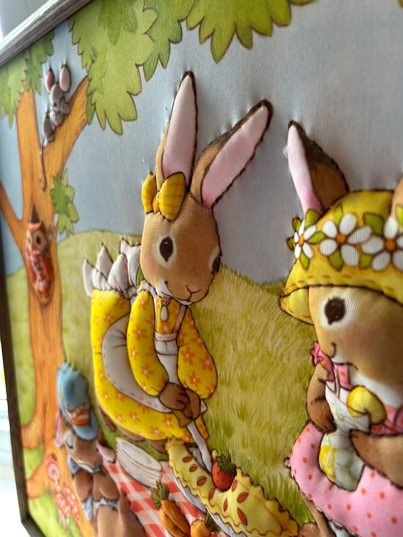 May include: A framed fabric wall art piece depicting two bunnies having a picnic in a forest setting. The bunnies are wearing dresses and have floral headbands. The scene includes a picnic basket, a pie, and a tree with a squirrel.