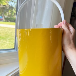 Può includere: Un secchiello per il ghiaccio cilindrico con una finitura gialla lucida e uniforme e un manico ad arco bianco liscio. Il contenitore presenta un pomello rettangolare trasparente sul coperchio, offrendo un design semplice e funzionale.