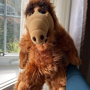 May include: A plush toy of the character Alf, featuring a tan snout, dark eyes, and shaggy brown fur. The toy is held up against a window with a curtain in the background. The toy's fur is a mix of brown and tan.