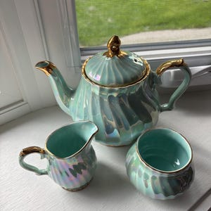 May include: A three-piece tea set comprising a teapot with a lid, a creamer, and a sugar bowl. Each piece is crafted from light green ceramic with a pearlescent, iridescent finish and is accented with shiny gold trim on the handles and rims.