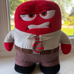 May include: A plush toy character with a bright red, square-shaped head, an angry facial expression, and embroidered eyes. The figure wears a short-sleeved white collared shirt, a red and green patterned necktie, brown trousers, and black shoes.