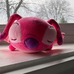 May include: A plush toy in the shape of a pink alien with large ears and purple accents. The toy has closed eyes and is resting on a white surface. The background shows a window with a view of bare trees.