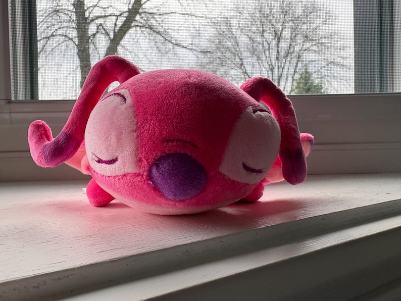 May include: A plush toy in the shape of a pink alien with large ears and purple accents. The toy has closed eyes and is resting on a white surface. The background shows a window with a view of bare trees.