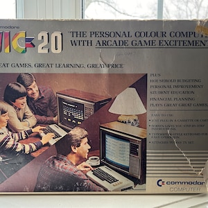 Può includere: Una scatola vintage per un computer personale a colori Commodore VIC-20. La scatola presenta una foto di tre persone che usano il computer e una descrizione delle sue funzionalità, tra cui la pianificazione familiare, il miglioramento personale, l'istruzione degli studenti, la pianificazione finanziaria e i giochi arcade. Il testo sulla scatola recita "Il computer personale a colori con l'eccitazione dei giochi arcade."