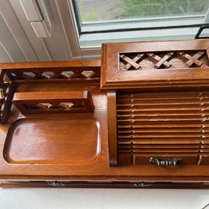 May include: A vintage wooden desk organizer with multiple compartments. It features a tray, a small drawer, and a slatted roll-top section. The organizer has decorative cutouts and a handle on top. The wood has a warm, medium-brown tone.