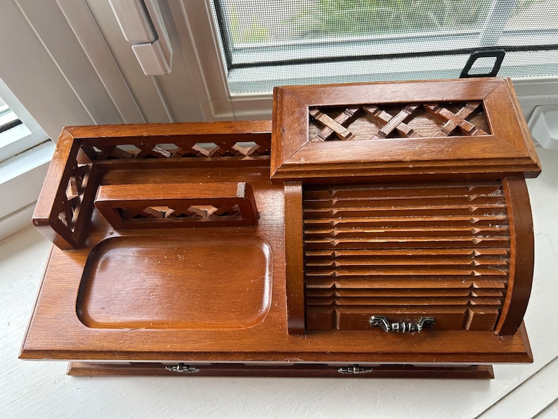 May include: A vintage wooden desk organizer with multiple compartments. It features a tray, a small drawer, and a slatted roll-top section. The organizer has decorative cutouts and a handle on top. The wood has a warm, medium-brown tone.