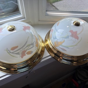 May include: Two dome-shaped ceiling light fittings featuring white glass shades with a floral pattern in shades of pink, orange, and green. Each fitting has a polished gold-tone metal base and a matching gold-tone decorative knob at the centre top.