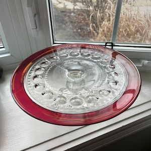 May include: A clear glass cake stand with a raised, textured centre and a wide, red rim. The stand is designed for serving desserts or snacks.