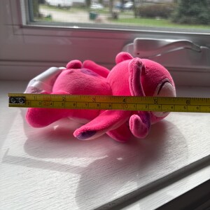 May include: A bright pink plush toy bunny, approximately 6 inches long, with purple accents on its ears and feet. The bunny is lying on a white surface near a window, with a measuring tape alongside it.