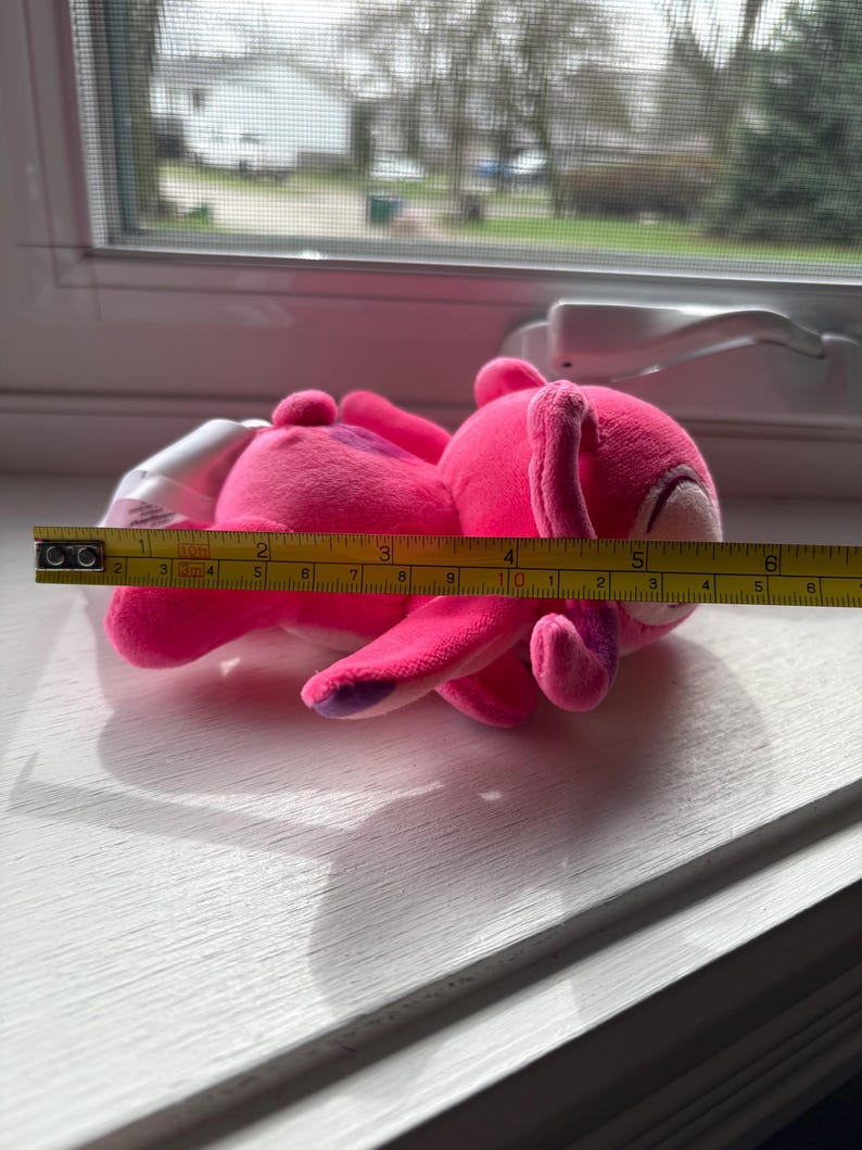 May include: A bright pink plush toy bunny, approximately 6 inches long, with purple accents on its ears and feet. The bunny is lying on a white surface near a window, with a measuring tape alongside it.