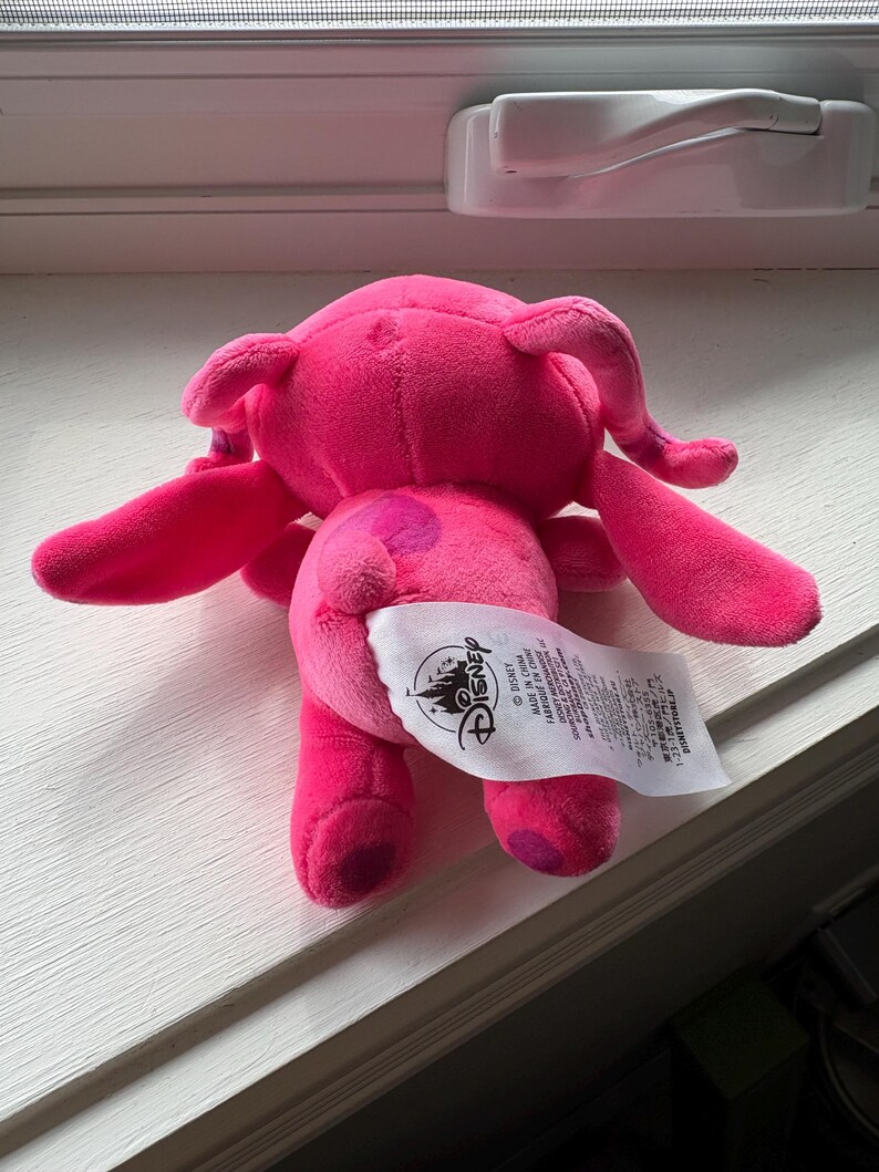 May include: A plush pink Angel doll, featuring large ears, long arms, and a tail. The doll has a Disney tag attached. The plush toy is made of soft material and is designed to resemble the character Angel.
