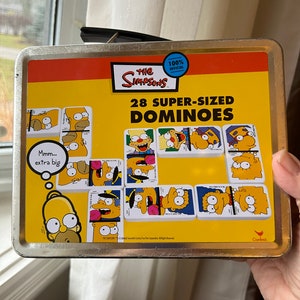May include: A metal tin box with a yellow background and the words "The Simpsons 100% Official 28 Super-Sized Dominoes" printed on it. The box features images of characters from the Simpsons cartoon, including Homer, Marge, Bart, Lisa, and Maggie. The box is closed and has a handle on the top.