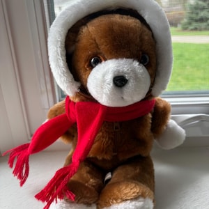May include: A plush teddy bear with brown fur, a white muzzle, and white paw pads. The bear wears a brown hooded jacket with a white faux fur trim around the hood and a bright red scarf with fringed ends. The jacket features a visible front zip.