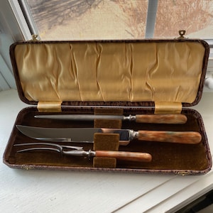 May include: A vintage carving set in a brown case. The set includes a carving knife, fork, and sharpening steel, each with a light brown and cream-colored handle. The case has a gold satin lining.