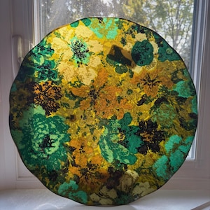 May include: A circular decorative item with an abstract floral pattern. It displays a blend of yellow, green, and brown hues, giving it a lively, textured look. The edges are irregular, enhancing its distinctive appeal. The diameter is approximately 30 cm.