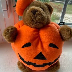 May include: A brown teddy bear wearing a plush orange pumpkin costume with a carved jack-o'-lantern face. The bear is sitting on a white surface.