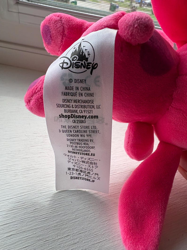 May include: A pink plush toy with a Disney label attached. The label includes the Disney logo, the text "MADE IN CHINA", and the shopDisney.com website address. The toy is a vibrant pink color.