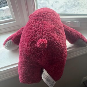 May include: A plush, magenta-colored bear toy with a fluffy texture. The bear has small arms and gray paws. A small, round tail is visible. A white tag is attached to the bear's bottom.