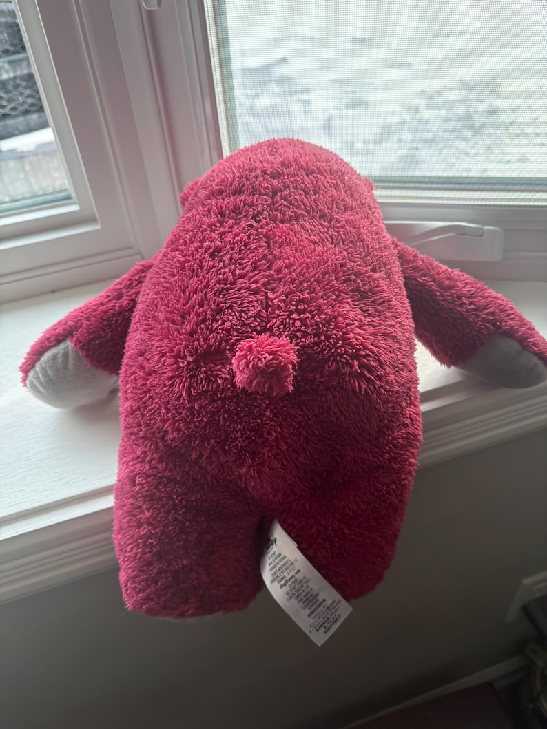 May include: A plush, magenta-colored bear toy with a fluffy texture. The bear has small arms and gray paws. A small, round tail is visible. A white tag is attached to the bear's bottom.