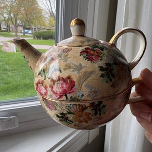 May include: A vintage tea-for-one set featuring a floral pattern with pink, red, and yellow flowers on a cream background. The teapot has a gold-colored handle and a matching lid with a gold finial. The teacup is stacked beneath the teapot.