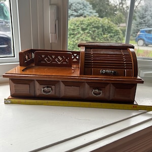May include: A vintage wooden jewelry box with a roll-top design and three small drawers. The box has a dark brown finish and silver-toned drawer pulls. A measuring tape is placed in front of the box. The box measures approximately 10 inches in length.
