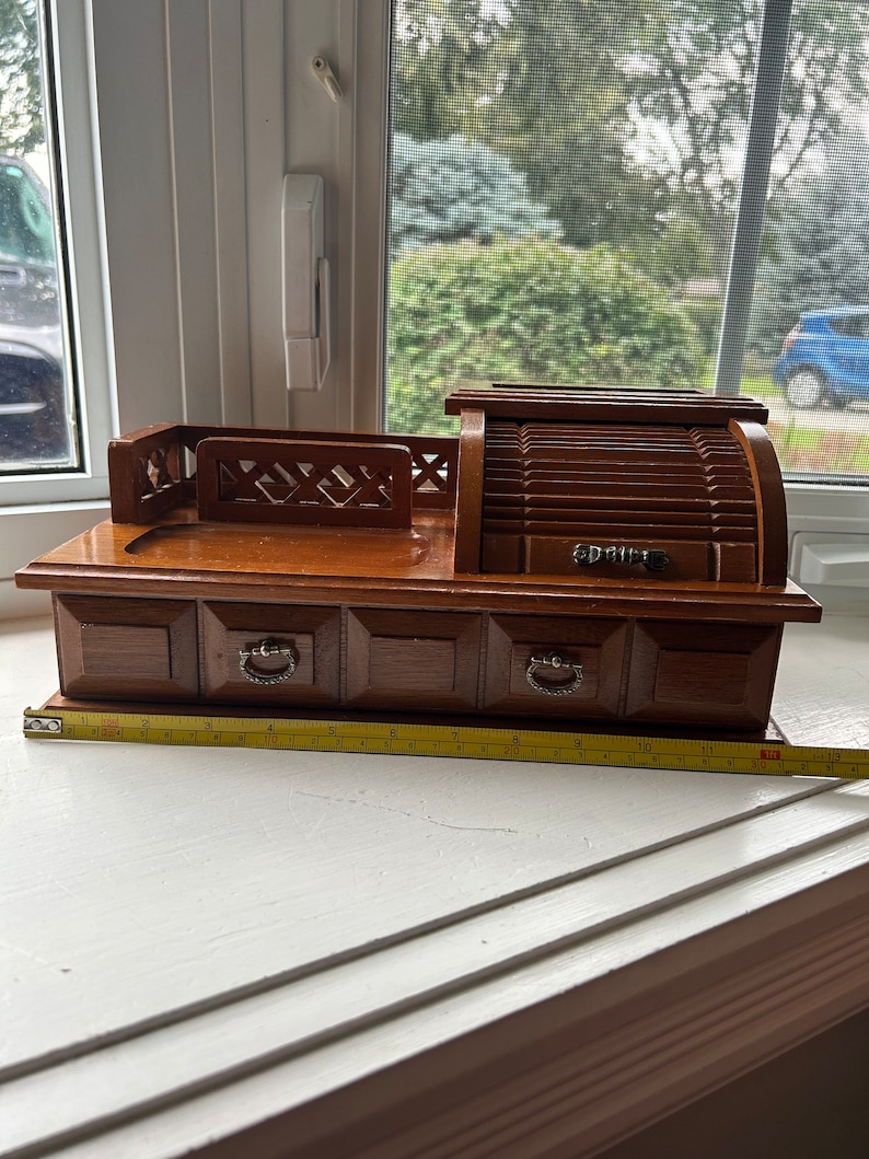 May include: A vintage wooden jewelry box with a roll-top design and three small drawers. The box has a dark brown finish and silver-toned drawer pulls. A measuring tape is placed in front of the box. The box measures approximately 10 inches in length.