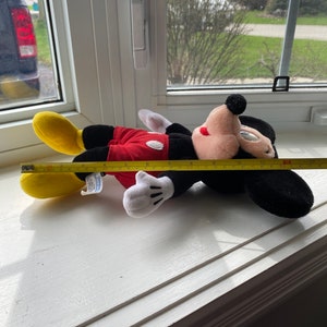 May include: A red and black plush Mickey Mouse toy lying on a white windowsill. The toy is approximately 12 inches long and has a yellow shoe on one foot. A yellow measuring tape is partially visible in the image.