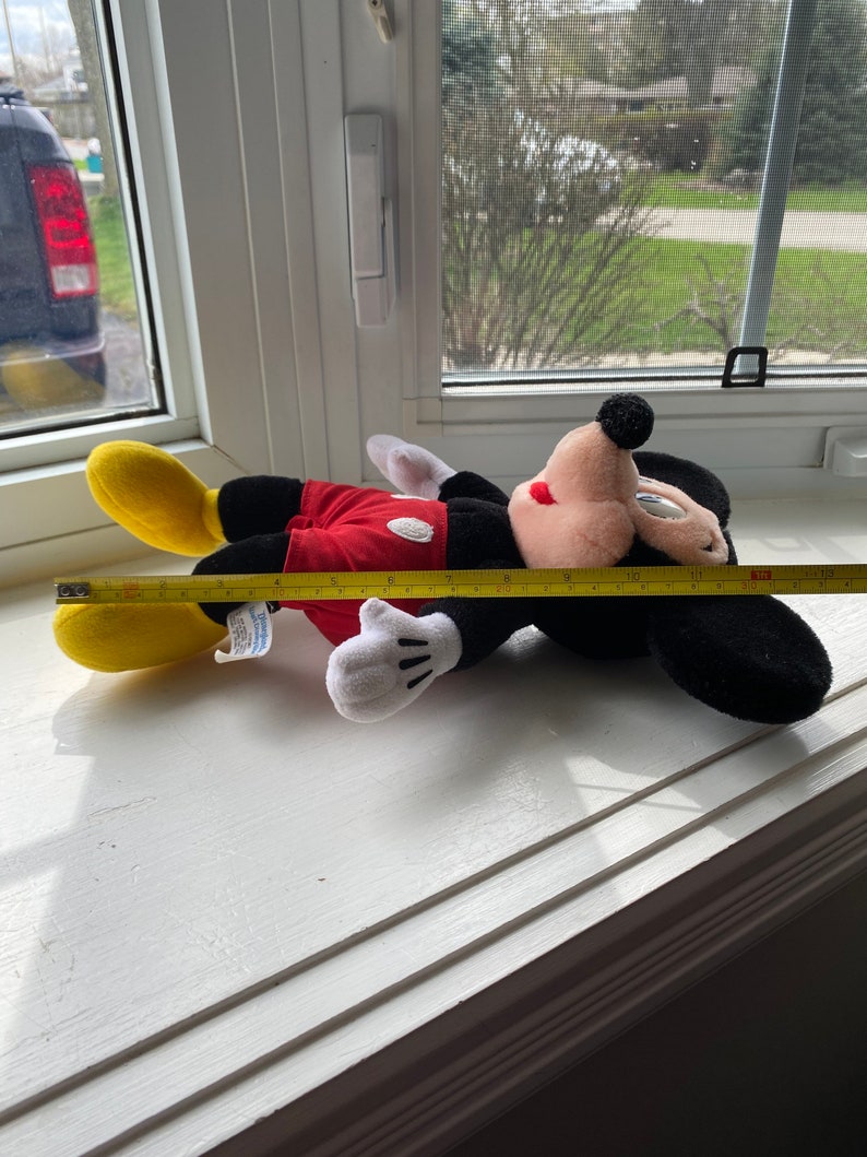 May include: A red and black plush Mickey Mouse toy lying on a white windowsill. The toy is approximately 12 inches long and has a yellow shoe on one foot. A yellow measuring tape is partially visible in the image.