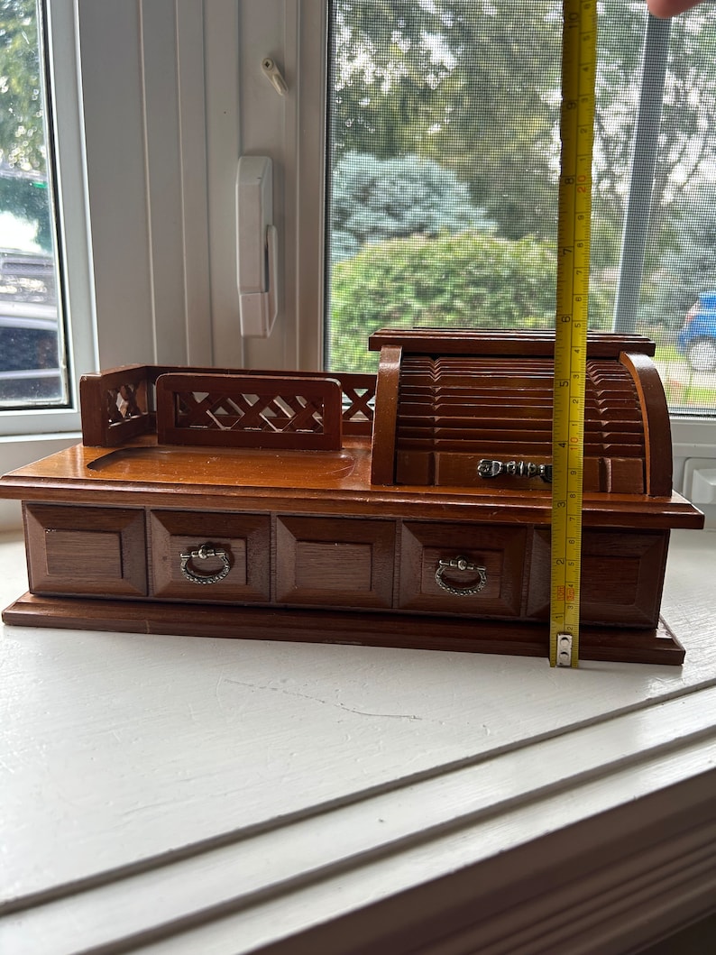 May include: A vintage wooden jewelry box with a roll-top design and four drawers. The box is approximately 9 inches tall. The drawers have metal pulls. The box is made of dark brown wood and sits on a white surface.