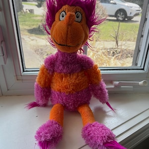 May include: A plush toy character with orange skin, a large nose, and expressive eyes. It has a pink and orange striped body, pink feet, and a vibrant pink, spiky hair. The toy is sitting on a white windowsill.