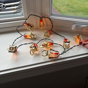 May include: A string of decorative lights featuring small, gold-colored lantern-shaped bulbs with red and orange accents. The lanterns are strung on a dark green wire and are arranged on a white windowsill. The lights are turned on, emitting a warm glow.