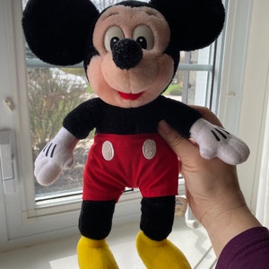 May include: A black and red plush Mickey Mouse toy with yellow shoes. The toy is standing on a white surface.
