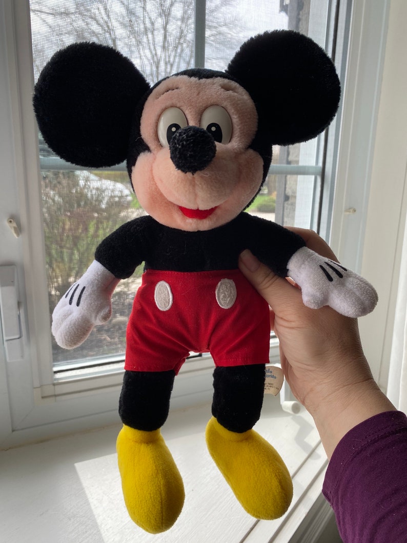 May include: A black and red plush Mickey Mouse toy with yellow shoes. The toy is standing on a white surface.