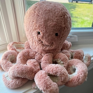 May include: A plush octopus toy featuring a soft, dusty pink textured exterior and a contrasting cream-colored underside on its eight curled tentacles. The stuffed animal has two small, dark, round eyes and a simple stitched mouth on its head.