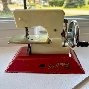 May include: A vintage Sew-O-Matic Junior sewing machine featuring a cream-colored upper body and a bright red metal base. The machine includes a silver-toned hand crank wheel with a black handle, a metal needle plate, and a needle assembly.