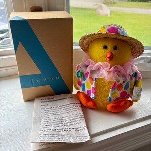 May include: A yellow plush chick figurine wearing a straw hat with a pink flower, a pink ruffled collar, and a vest with colourful polka dots. It is accompanied by a brown cardboard box labelled AVON and a white paper instruction sheet.