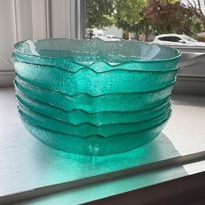 May include: A stack of six turquoise glass bowls with a textured, leaf-like pattern. The bowls have a wavy rim and are stacked on a white windowsill. The bowls are translucent, allowing light to pass through.