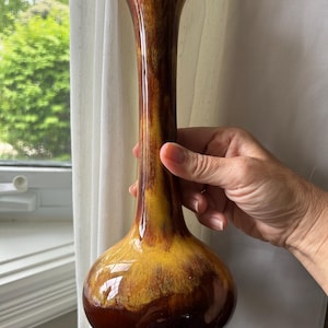 May include: A tall, slender vase with a bulbous base and flared top. The ceramic vase has a glossy finish with a blend of brown, yellow, and orange hues. The vase is held by a person's hand.