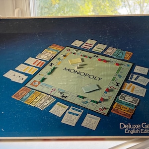 May include: A Monopoly Deluxe Game English Edition board game box featuring a dark blue background with a central game board layout. The board displays property spaces, Chance and Community Chest cards, paper money, and small green plastic houses.