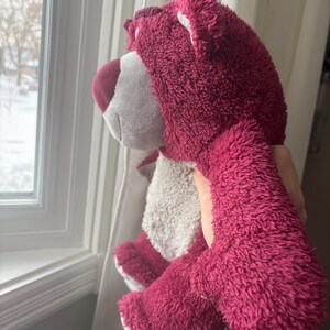 May include: A plush, magenta-colored teddy bear with a white muzzle and belly. The bear has a soft, fuzzy texture and is being held up near a window. The bear's ears and paws are white, and its nose is a darker shade of magenta.