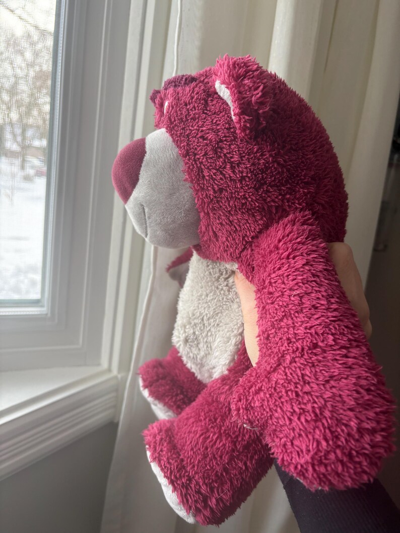 May include: A plush, magenta-colored teddy bear with a white muzzle and belly. The bear has a soft, fuzzy texture and is being held up near a window. The bear's ears and paws are white, and its nose is a darker shade of magenta.