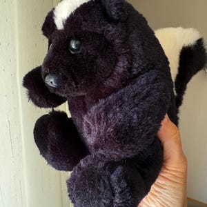 May include: A plush skunk toy featuring soft black fur with a prominent white stripe running from the top of its head down to its back. The stuffed animal has dark, round eyes, a small black nose, and a bushy white tail extending outward.