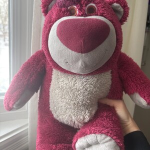 May include: A plush, pink teddy bear with a heart-shaped nose and a white belly. The bear has a friendly expression and is made of soft, fuzzy material. The bear is being held up in front of a window.