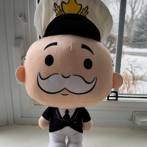 May include: A plush toy of a cartoon character wearing a white captain's hat with a gold star and a black and white uniform. The character has a large head, a mustache, and a friendly expression.