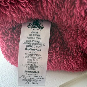 May include: Close-up of a plush, burgundy-colored fabric with a Disney label attached. The label includes the Disney logo, manufacturing details, and website information. The fabric appears soft and textured.